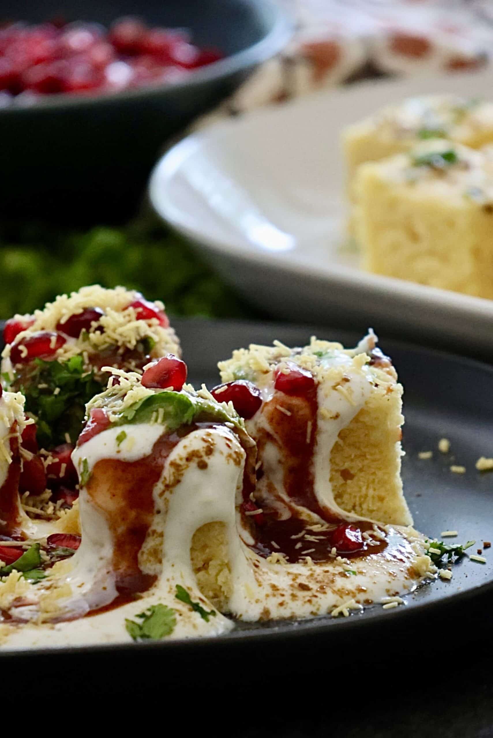 Overhead flatlay of khaman chaat served on a black plate with spiced yogurt, green chutney, tamarind chutney, sev, pomegranate arils and cilantro.