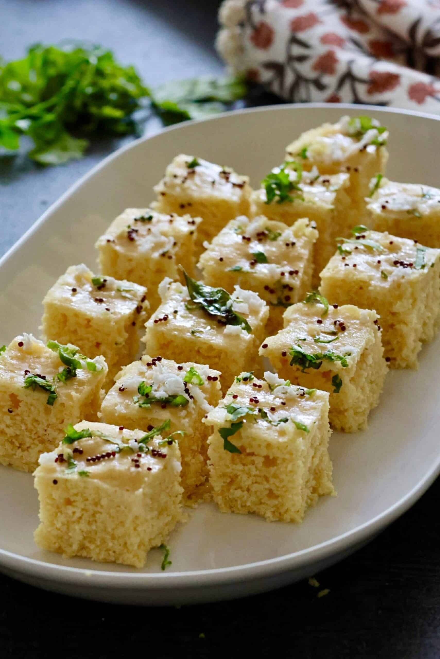 Freshly steamed Khaman cut into squares plated on a white plate, garnished with a curry leave and mustard seed tempering.