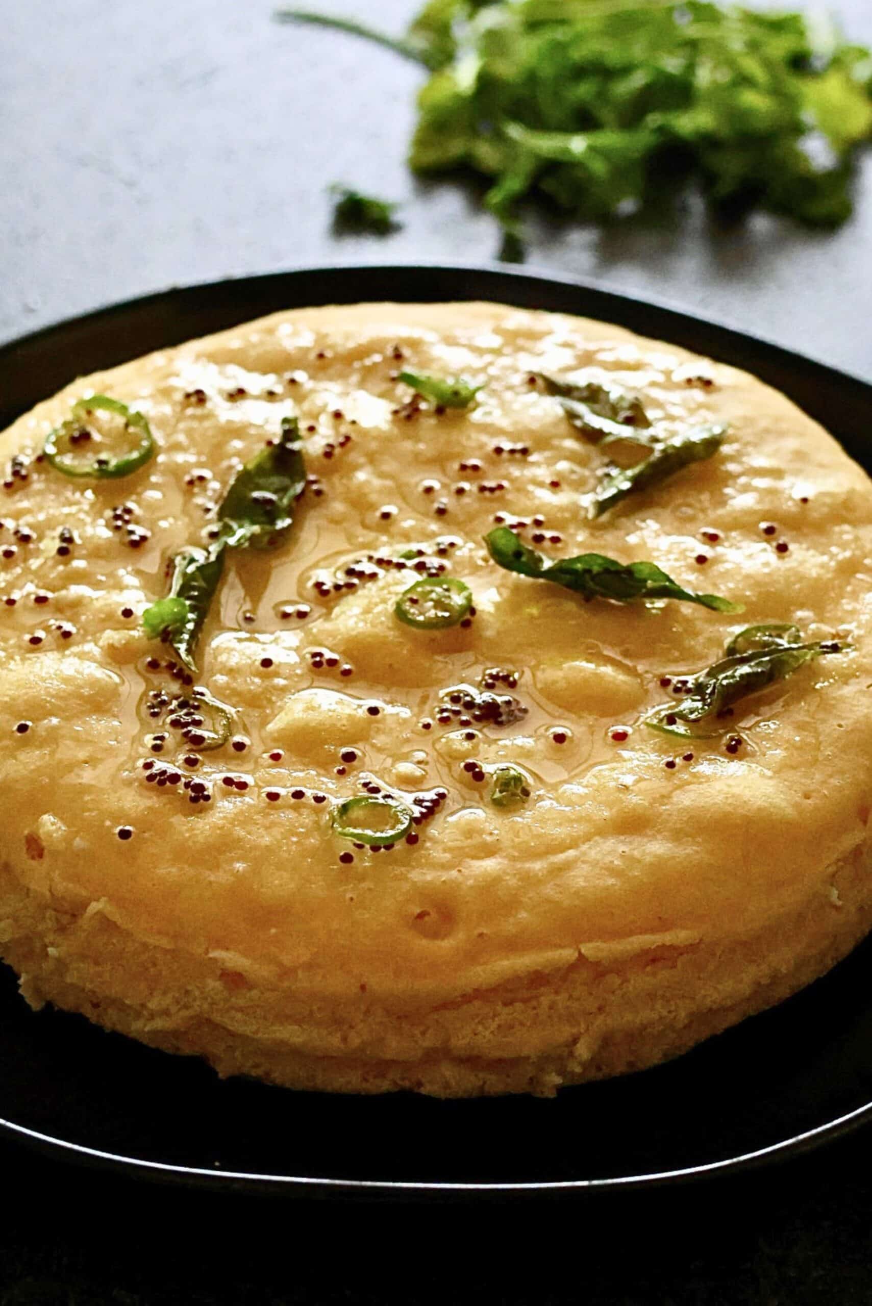 Whole steamed khaman dhokla cake fresh out of the Instant Pot in a round pan, golden yellow with a smooth tempering before cutting & tempering